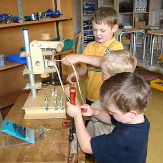 Trois enfants jouent ensemble autour d'une table de travail, manipulant un appareil en bois pour apprendre à jongler. L'un des enfants est en train de tirer sur une corde, tandis que les deux autres s'occupent de différents outils en bois, l'un tenant un 