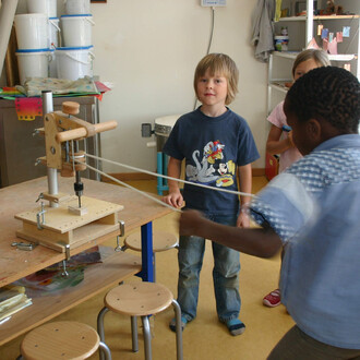 Un enfant avec des cheveux blonds et un t-shirt bleu se tient devant une machine de jonglerie en bois. Deux autres enfants sont visibles en arrière-plan. La machine, avec un mécanisme en bois et des cordes, est placée sur une table en bois avec quelques t