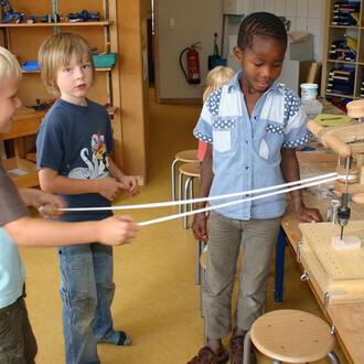 Enfants jouant ensemble dans une salle de classe, utilisant des cordes sur un matériel de jonglerie. Un enfant avec une chemise à motifs observe pendant que deux autres tiennent les cordes. Des chaises en bois et des outils de jonglerie sont visibles en a