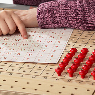 Main tenant une feuille de jeu avec des chiffres imprimés en rouge, positionnée sur une table en bois. À côté, il y a une rangée de pions rouges placés sur un fond en bois perforé.