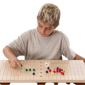 Un enfant blonde portant un t-shirt beige joue sur une table en bois perforée, disposant des billes colorées sur un plateau de jeux. Il regarde attentivement en déplaçant une bille verte. À côté, un dé est visible sur la table.