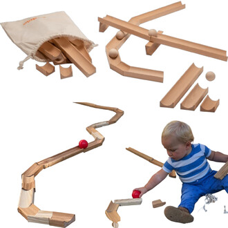 Ensemble de blocs en bois pour créer des parcours, comprenant des rampes et des billes. Un enfant assis interagissant avec des pièces, tandis qu'une balle rouge roule sur une structure en bois. Sac en tissu contenant des blocs en hauteur.