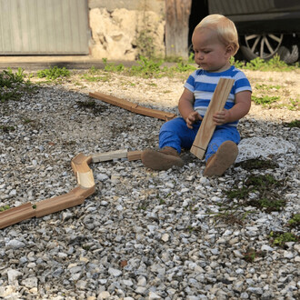 Un enfant assis sur un sol en gravier, jouant avec des éléments en bois. Il porte un t-shirt rayé bleu et blanc et des pantalons bleus. Devant lui, des morceaux de bois sont disposés pour former une structure. En arrière-plan, on peut voir un mur en pierr