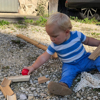 Un jeune enfant assis sur un sol en gravier, jouant avec une balle rouge. Il porte un t-shirt rayé bleu et blanc et un pantalon bleu. Autour de lui, des morceaux de bois de construction sont dispersés, formant une structure incomplète. En arrière-plan, on