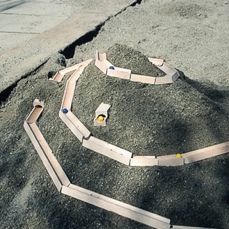 Piste de billes en bois disposée sur un monticule de sable, avec des billes de différentes couleurs (jaune et bleue) roulant dans les canaux.