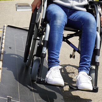 Personne assise dans un fauteuil roulant, en train d'utiliser une rampe d'accès, avec des jambes croisées et des baskets blanches. Le fauteuil est en position inclinée sur la rampe.