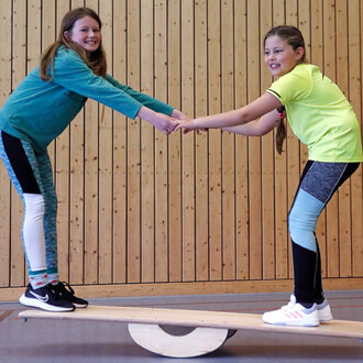 Deux filles s'amusent sur un équilibrateur en bois, se tenant par les mains pour conserver l'équilibre. L'une porte un sweat turquoise et un legging noir et blanc, tandis que l'autre est vêtue d'un t-shirt jaune et d'un legging bleu et noir. Le fond est c