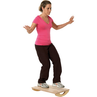 Donna con maglietta rosa e pantaloni neri, in equilibrio su una tavola di legno, con le braccia tese per mantenere l'equilibrio.