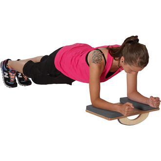 Femme en position de planche sur un accessoire d'entraînement, portant un haut rose et un pantalon noir. Elle utilise ses avant-bras pour se soutenir sur la plateforme, le visage concentré. Des chaussures de sport noires et colorées complètent sa tenue.