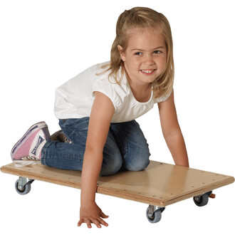 Fille assise sur une planche en bois avec roulettes, souriante, portant un tee-shirt blanc et un jean. Elle est positionnée à genoux sur la planche, avec les mains posées dessus.