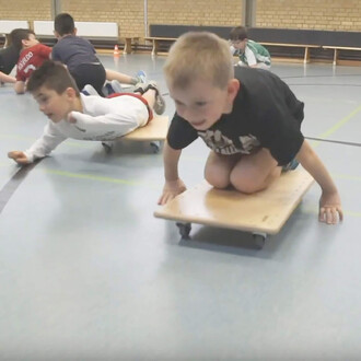 Deux enfants s'amusent sur des planches à roulettes dans une salle de sport. L'un est allongé à plat ventre sur sa planche, tandis que l'autre est en position accroupie, souriant. D'autres enfants jouent en arrière-plan. Le sol est en linoléum avec des li