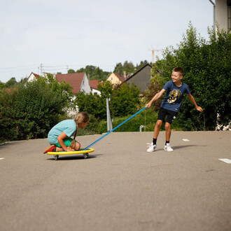 Deux enfants jouent en extérieur sur une route. Un garçon, souriant et debout, tire une fille assise sur un skate avec une corde bleue. La fille porte un t-shirt turquoise et se tient en avant, tandis que le garçon porte un t-shirt bleu avec un motif et u