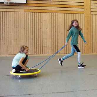 Deux enfants jouent ensemble dans une salle de sport. L'un est assis sur un chariot roulant jaune, tiré par l'autre enfant qui court en riant. Le sol est en linoléum et les murs sont en bois.