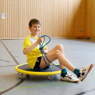 Un enfant souriant porte un t-shirt jaune et est assis sur une planche à roulettes, tenant une corde bleue. Il est dans une salle de sport avec des murs en bois et un sol marqué par des lignes de jeu.