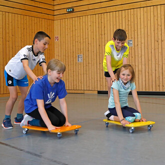 Quatre enfants jouent dans une salle de sport. Deux d'entre eux sont debout en arrière, poussant les deux autres qui sont assis sur des planches roulantes orange et jaune. Les enfants sourient et semblent s'amuser. La salle est dotée de murs en bois et d'