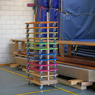 Étagère de rangement en bois avec plusieurs étagères de couleurs vives (orange, jaune, vert, bleu, rose) et munie de roulettes, installée dans une salle de gym avec des bancs de gym en arrière-plan.