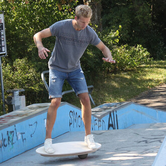 Jeune homme en t-shirt gris et shorts, en train de jongler sur une planche d'équilibre sur une rampe de skate, avec un décor en arrière-plan comprenant des arbres et des graffitis sur les murs.