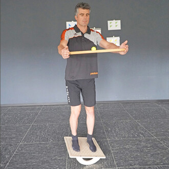 Homme en tenue de sport se tenant debout sur un équipement de jonglerie, avec les yeux fermés, tenant un bâton horizontalement et faisant léviter une balle en plastique sur celui-ci. Arrière-plan de couleur sombre avec des illustrations accrochées au mur.