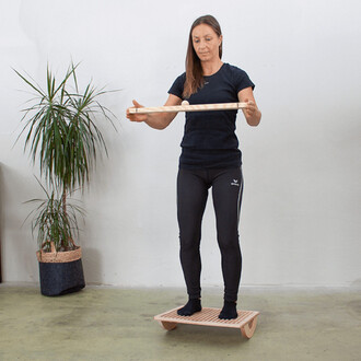 Femme debout sur une plateforme en bois, tenant une barre au-dessus d'elle avec une boule. Elle est habillée en noir, avec des leggings ajustés et un t-shirt. En arrière-plan, une plante verte et un pot tressé.