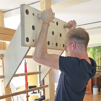 Homme effectuant un exercice de traction sur une planche d'escalade en bois Pedalo®, fixée à une structure de gymnastique. La planche présente plusieurs poignées et trous pour un entraînement varié. En arrière-plan, on aperçoit une salle d'activités avec 