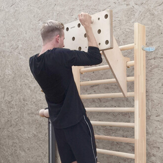 Un homme s'entraîne sur une planche d'escalade Pedalo® fixée à une structure. Il est de dos, tenant des poignées en bois. La planche présente des trous ronds pour l'escalade, et des barres horizontales en bois sont visibles en dessous. Le fond est constit