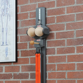 Support mural de jonglerie avec deux boules en bois et une barre en bois, fixé sur un mur en briques. Des bandes élastiques orange sont intégrées sous l'appareil. Un schéma anatomique est visible en arrière-plan.