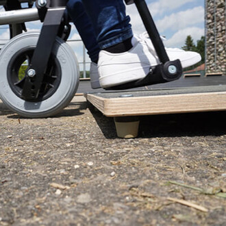 Roues d'un équipement de jonglerie sur un sol extérieur, avec un pied portant une chaussure blanche qui repose sur une surface en bois. La photo montre le contact entre les roues et la plateforme.