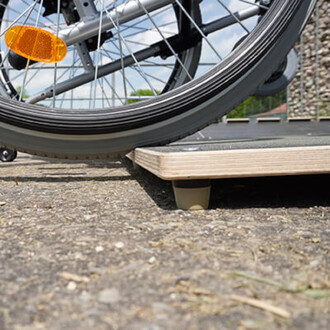 Vue en gros plan d'une roue de vélo, avec un réflecteur orange visible, posée sur une plateforme en bois sur un sol en gravier. Le fond montre un ciel partiellement nuageux et un espace ouvert.