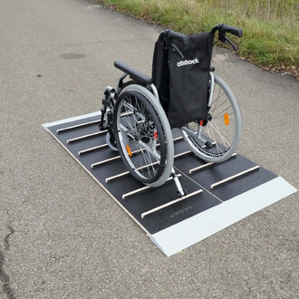 Tapis de déplacement en plastique pour fauteuil roulant avec des lattes en bois, posé sur une route. Le fauteuil roulant est garé sur le tapis, qui est conçu pour faciliter le passage sur des surfaces irrégulières.