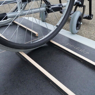 Roue d'un fauteuil roulant sur une surface de jeu en bois, avec des bandes de matériel noir et des éléments en bois, mettant en avant le produit Pedalo.