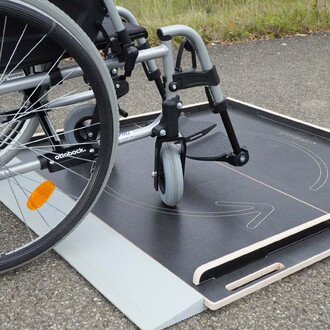 Rampe en bois pour fauteuil roulant, avec un fauteuil roulant partiellement posé dessus, sur un sol en asphalte. La rampe présente une surface noire texturée et des bords en bois naturel, avec un marquage indiquant une direction.