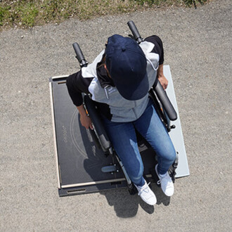 Vue aérienne d'une personne assise dans un fauteuil roulant sur uneplateforme, sur un chemin en gravier. La personne porte une casquette et un sweat à capuche, et a des jeans et des chaussures blanches.