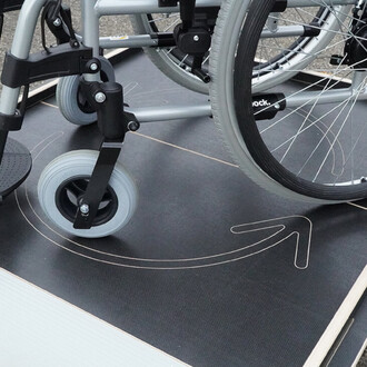 Roues d'un fauteuil roulant sur une surface noire avec un motif de flèche indiquant une direction. La photo met en avant des roulettes grises et la structure en métal du fauteuil.