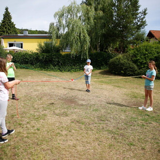 Quatre enfants jouent dans un jardin. Ils tiennent chacun une corde qui se croise au centre, où se trouve une boule rouge. L'un des enfants est au centre, vêtu d'un t-shirt blanc et d'une casquette, tandis que les autres sont disposés autour de lui, tenan