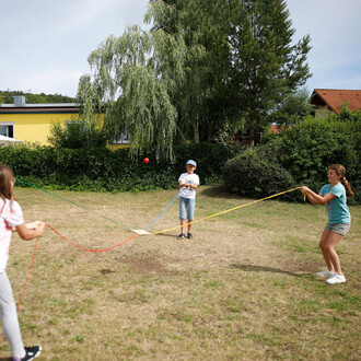 Trois enfants jouent en extérieure sur un terrain en herbe. Deux enfants tiennent des cordes de jonglerie de différentes couleurs, tandis qu'un troisième enfant au centre tente d'attraper une petite balle rouge qui est en l'air. En arrière-plan, se dresse