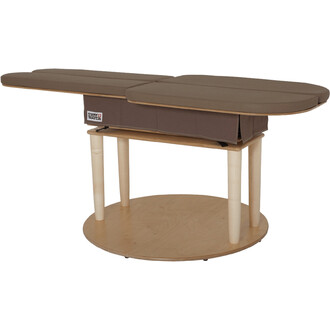 Table de massage en bois avec deux plateaux inclinés, recouverts de tissu brun. Elle possède une base en cercle, avec un espace de rangement sur la partie inférieure. Les pieds sont en bois clair.