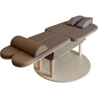 Table de massage en bois, avec un revêtement en similicuir marron, deux têtières et des repose-jambes arrondis. Elle repose sur un socle circulaire en bois.