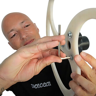 Un utilisateur démontrant le montage du Plug Hub Flowbonacci, en plaçant une pièce métallique à l'intérieur d'un cadre circulaire. La personne a la tête rasée et porte un t-shirt noir avec un logo. Le fond est blanc, mettant en évidence le matériel de jon