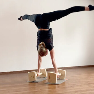 Personne effectuant un équilibre sur les mains sur des barres parallèles en bois, avec une posture acrobatique. Le sol est en bois, et le fond est neutre. La personne porte un haut noir et un legging noir, et a les cheveux attachés.