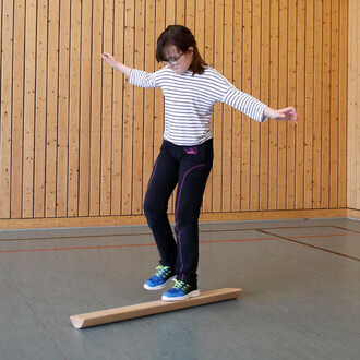 Une jeune fille se tient en équilibre sur une barre en bois dans une salle de gym, avec des murs en bois apparents. Elle porte un t-shirt rayé blanc et noir, ainsi que des pantalons noirs avec des accents violets. Ses bras sont légèrement écartés pour mai