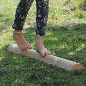 Enfant pieds nus équilibré sur une barre en bois, située sur une pelouse verte, portant un pantalon à motifs floraux.