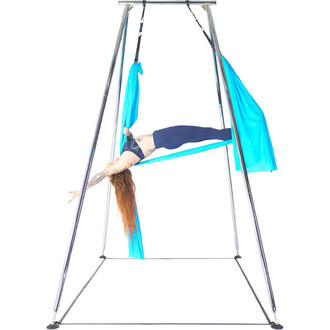 Femme en position de yoga suspendue, utilisant un portique avec des tissus aériens turquoise. Elle est allongée sur le dos, bras tendus vers l'avant, les jambes soutenues par le tissu. Le portique est en métal chromé, avec des structures solides, sur un f