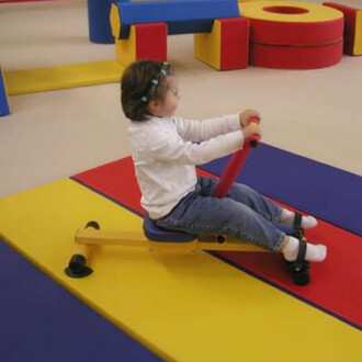 Un enfant assis sur un rameur pour enfants, tenant une barre rouge, sur un tapis coloré. Le fond montre des blocs de jeux en rouge, jaune et bleu.