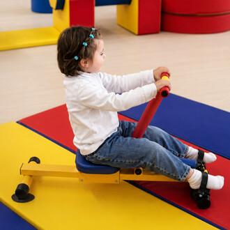 Ein Kind sitzt auf einem Kinderrudergerät mit gelbem Rahmen, blauem Sitz und roten Griffen, das auf einer bunten Matte auf einem Spielplatz steht.