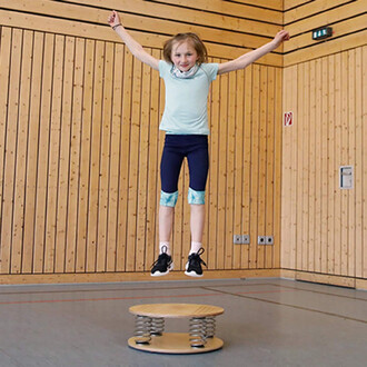 Une fille blonde saute avec les bras levés au-dessus d'un dispositif de saut en bois et ressorts, dans une salle de sport avec des murs en bois. Elle porte un t-shirt turquoise et des shorts sombres avec des motifs, ainsi que des baskets noires.