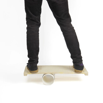 Jambes d'une personne en pantalon noir debout sur une plateforme en bois Rolla Bolla Status, la planche reposant sur des rouleaux blancs en PVC.