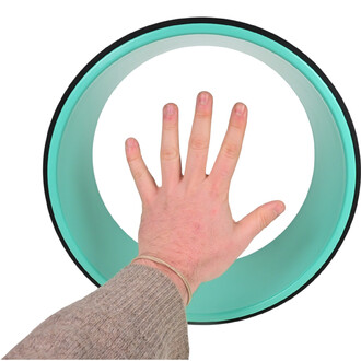 Main posée sur une roue de yoga de couleur verte, vue du dessus. La roue présente une bordure noire et un intérieur lisse. Le pouce et les doigts de la main sont visibles, ainsi qu'une partie du poignet avec un élastique.