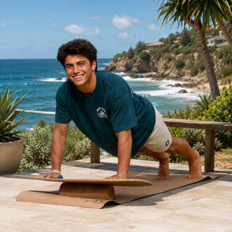 Homme souriant en tee-shirt bleu effectuant une posture de yoga sur une planche posée sur un rouleau de yoga en liège, en extérieur sur une terrasse avec vue sur la mer et des palmiers.