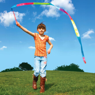 Petite fille courant sur l'herbe verte avec un ruban arc-en-ciel attaché à une baguette. Elle porte un t-shirt orange à motifs et un jean bleu. Le ciel est bleu avec des nuages blancs, créant une ambiance joyeuse et ensoleillée.