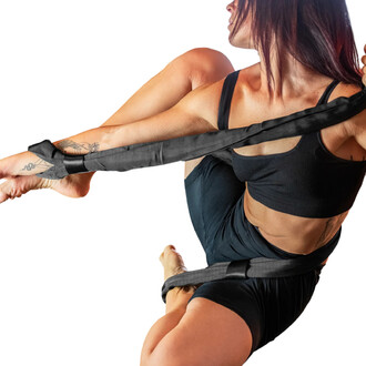 Athlète en position de suspension utilisant des sangles aériennes, portant un haut noir et un bas noir, avec des tatouages visibles sur le bras. Les sangles sont attachées à ses bras et cuisses, démontrant une technique de jonglerie. Fond blanc.